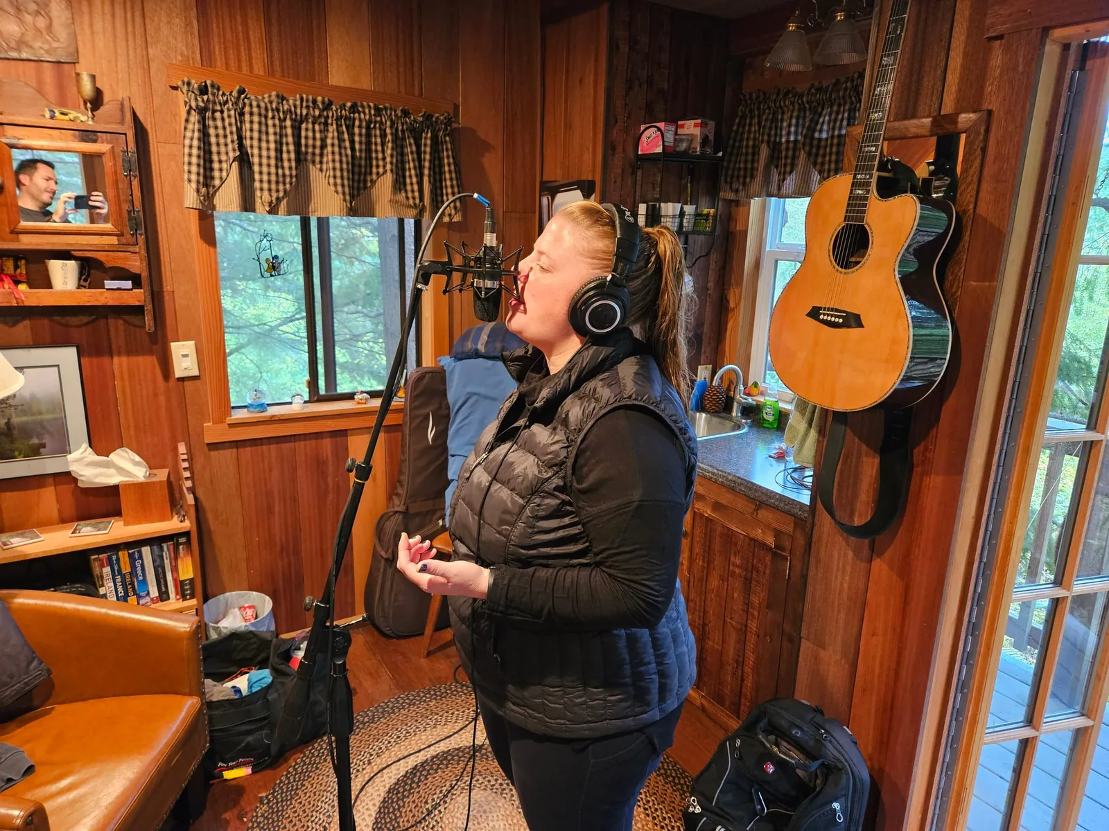 Our singer recording vocals in headphones at a studio mic in a cozy wood-paneled room, an acoustic guitar on the wall.
