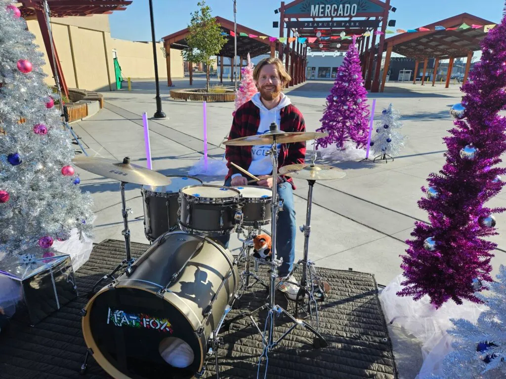 Our drummer grinning at the Fat Fox drum kit among purple, pink and silver Christmas trees, the Mercado sign in the distance.