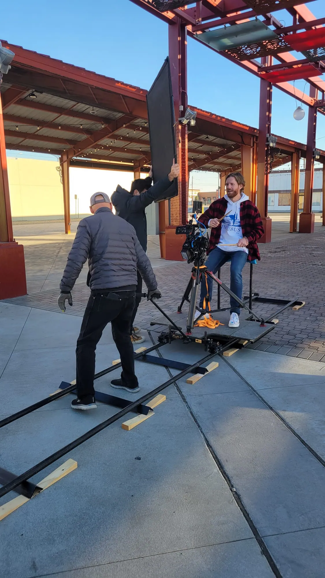 Our drummer, sticks in hand, filmed up close as the crew rolls a camera dolly on track and holds up a big black panel.