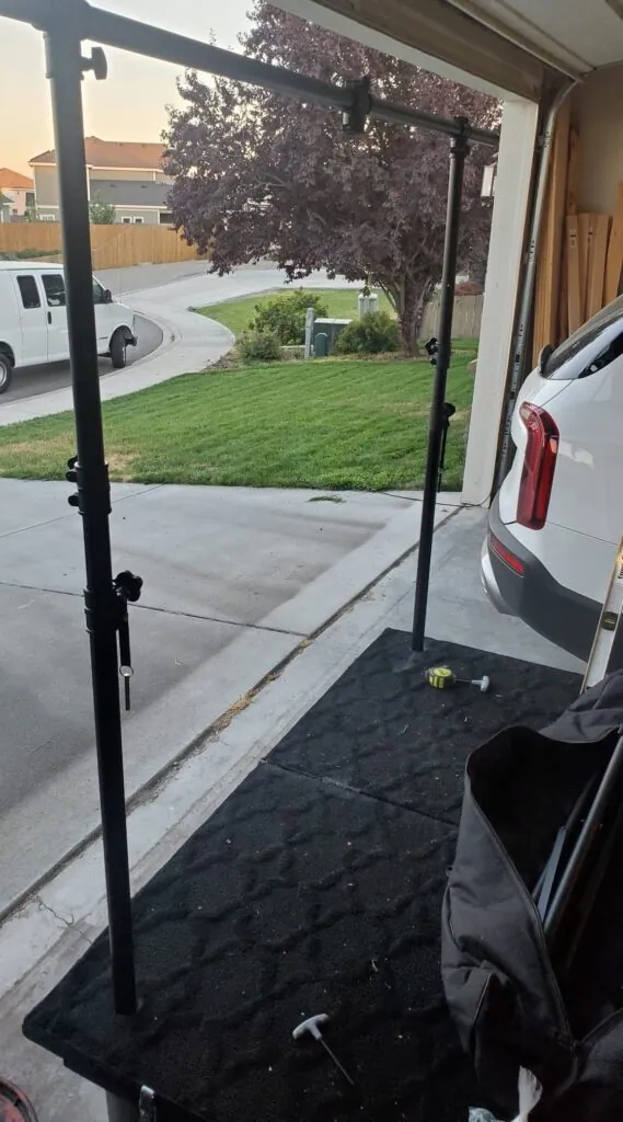 A tall black stand frame with a crossbar set up on a carpeted platform in the driveway, a tape measure nearby.