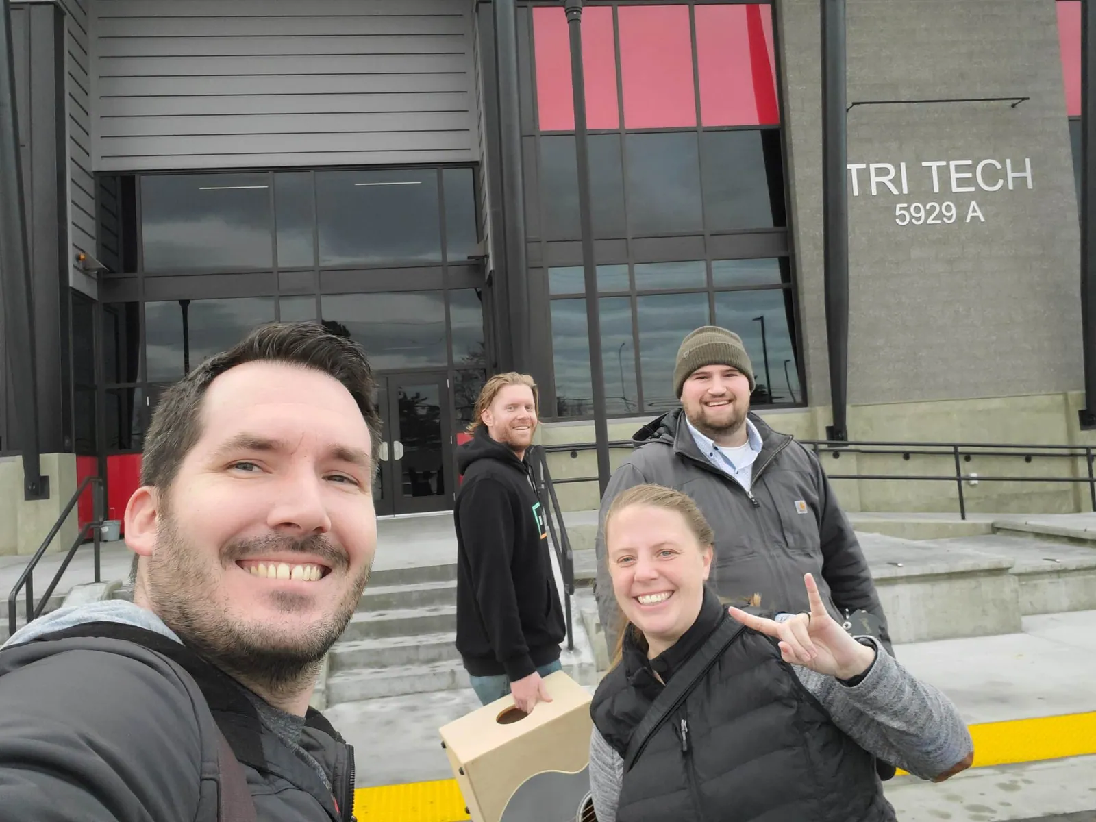 The four of us smiling for a selfie outside the Tri-Tech Skills Center, carrying a cajon and an acoustic guitar.