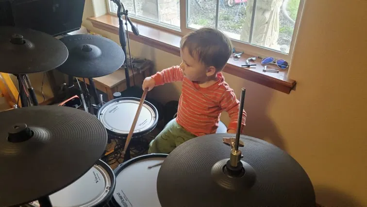 Little Lucas sitting at an electronic drum kit by the window, drumstick in hand.