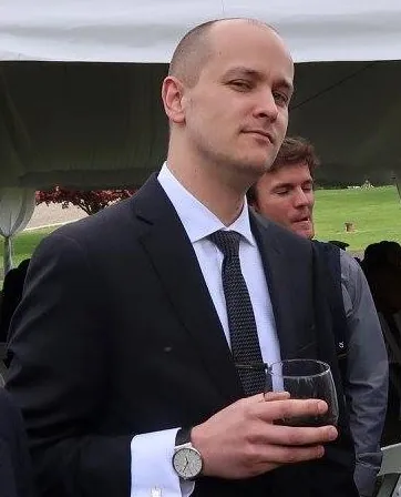 TJ giving a cool look in a dark suit and tie, a glass of red wine in hand, under a white tent outdoors.