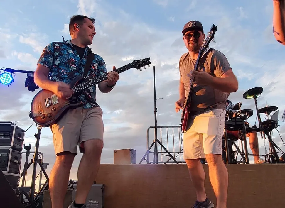 Two of us rocking out with our guitars on an outdoor stage at sunset, drums set up behind us.