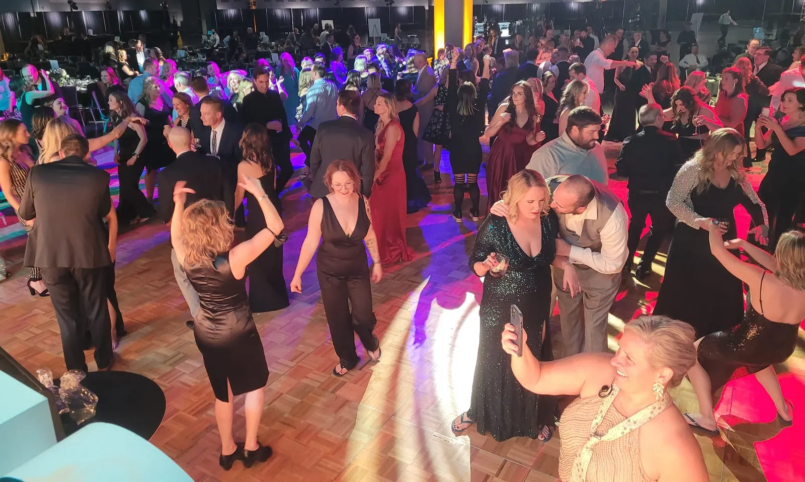 A packed dance floor of guests in evening wear, dancing under purple and gold lights