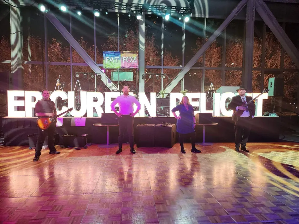 The four of us on an empty dance floor in front of giant lit-up letters spelling Epicurean Delight, our stage set up behind them