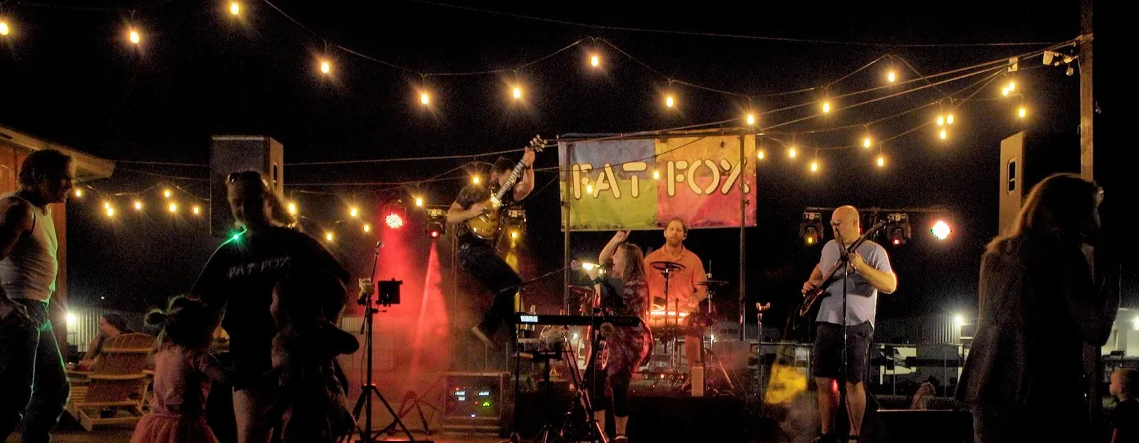 An outdoor party at night, our band on a small stage under string lights and our Fat Fox banner, our stage lights glowing red and green, people dancing in front