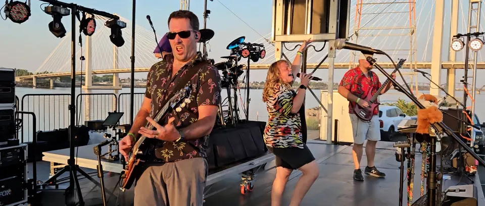 Our guitarist pulling a face mid-song while our singer dances behind him, on a riverside stage at sunset