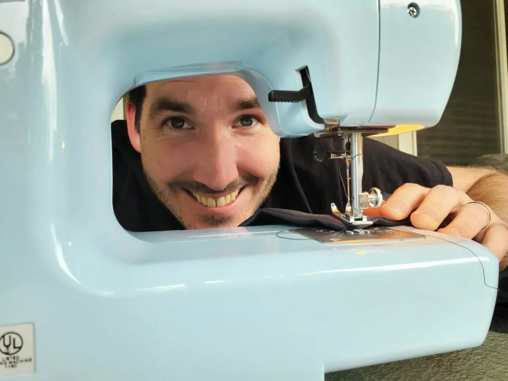 One of us grinning through the arm of a light blue sewing machine while stitching black fabric.