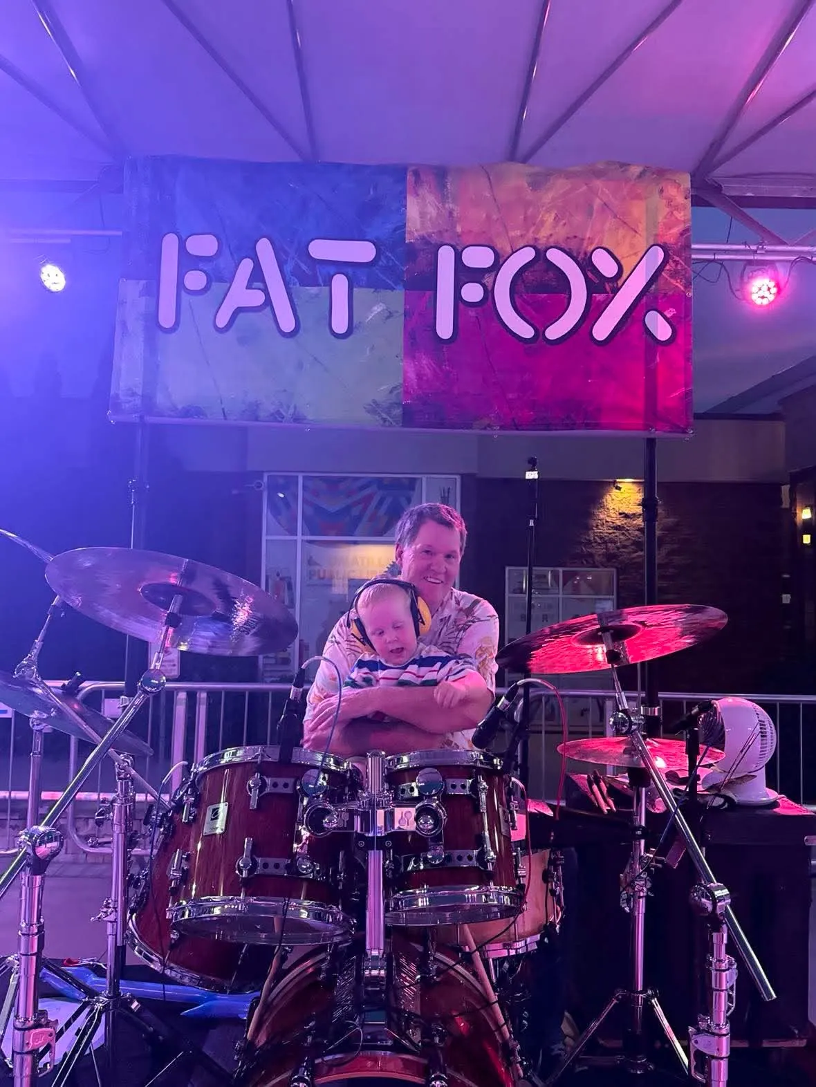 Steve behind the drum kit under the Fat Fox banner, with a little one in yellow ear protection on his lap