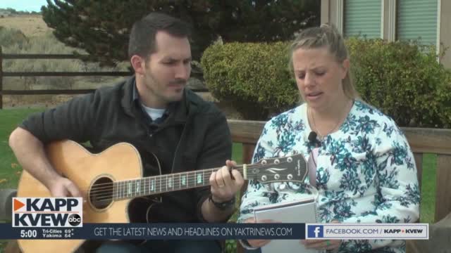 Kali singing her song on an outdoor bench, accompanied on acoustic guitar, in an ABC KAPP-KVEW news clip.